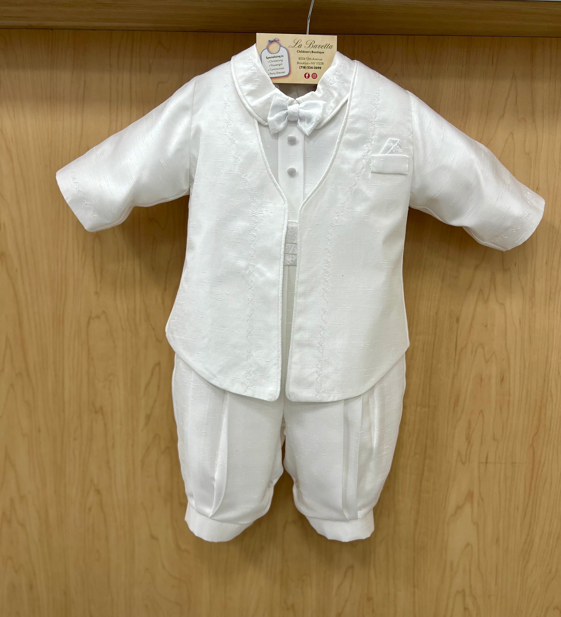 A white knee-length christening outfit with a jacket and long pleated pants, featuring a cumberbund, suspender, and bowtie. The outfit is displayed on a wooden hanger.