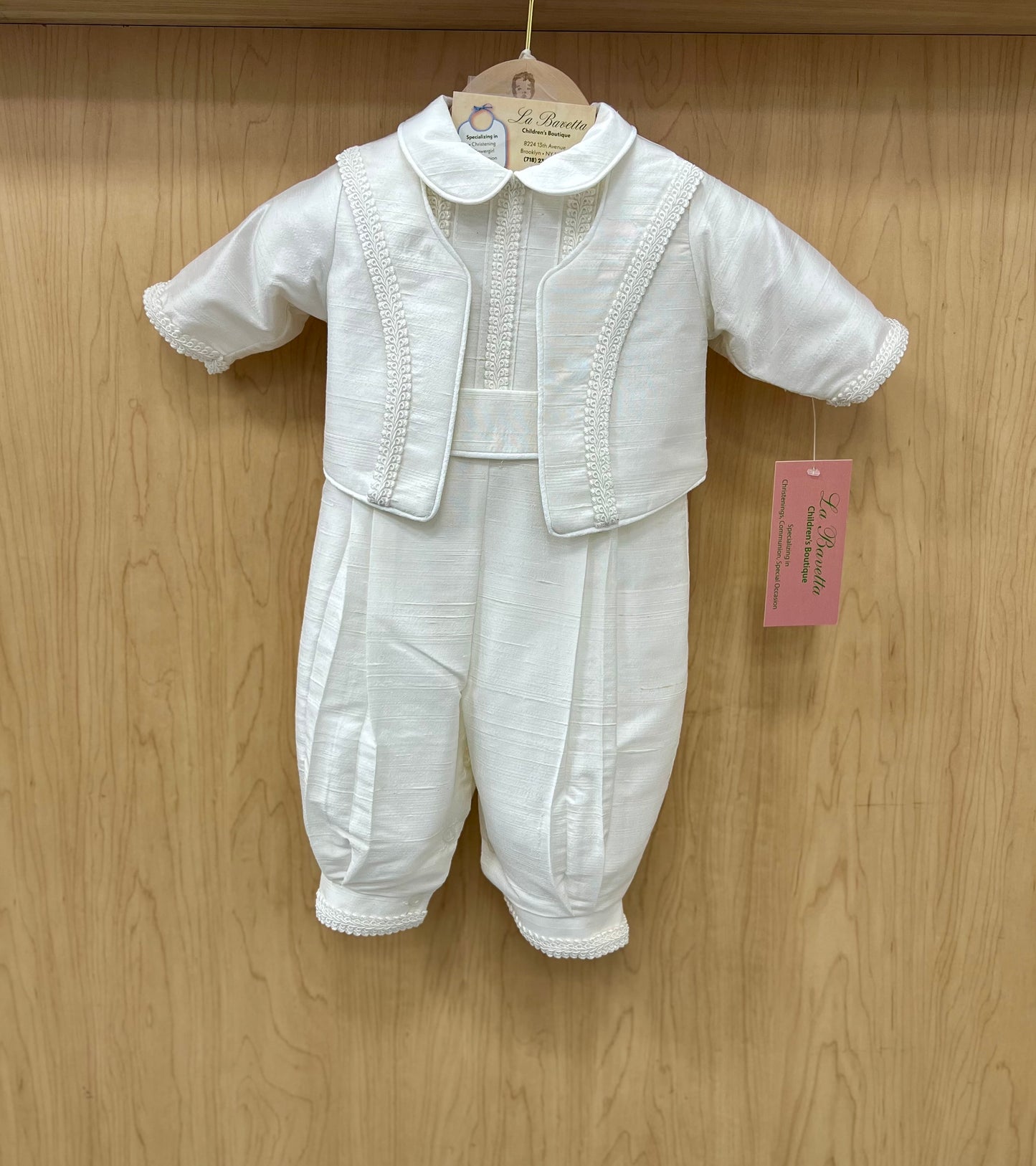 A white silk christening suit for a baby boy, with embellishments such as cording and piping, displayed on a wooden hanger.