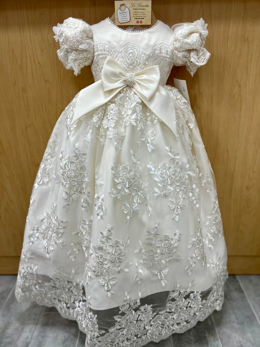 A beige silk baptism wear dress for a baby girl, featuring 3D lace, beaded details, and a large bow at the waist.