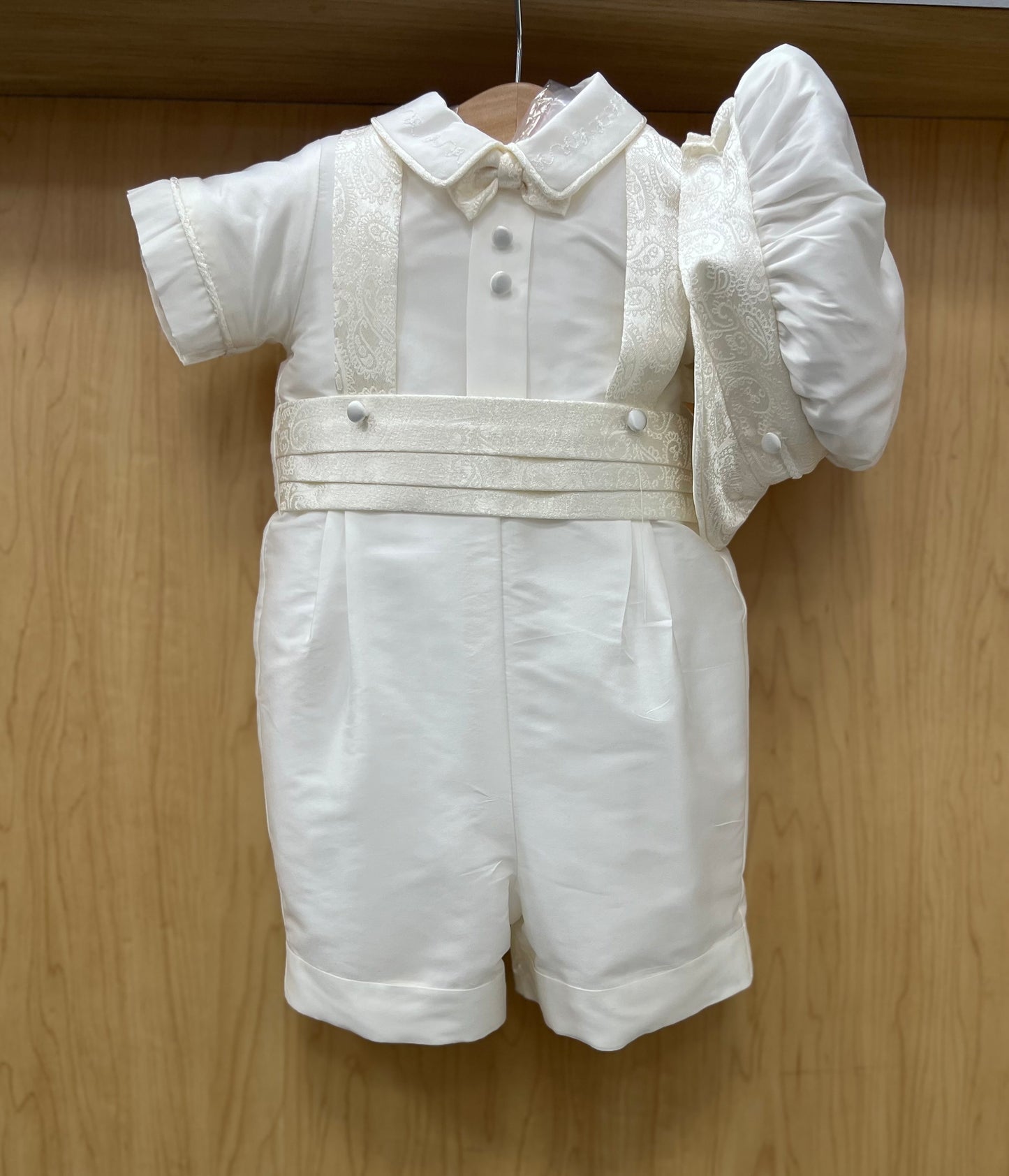 White silk baby shorts christening outfit with jacquard suspenders and a flat cap, displayed on a wooden hanger.