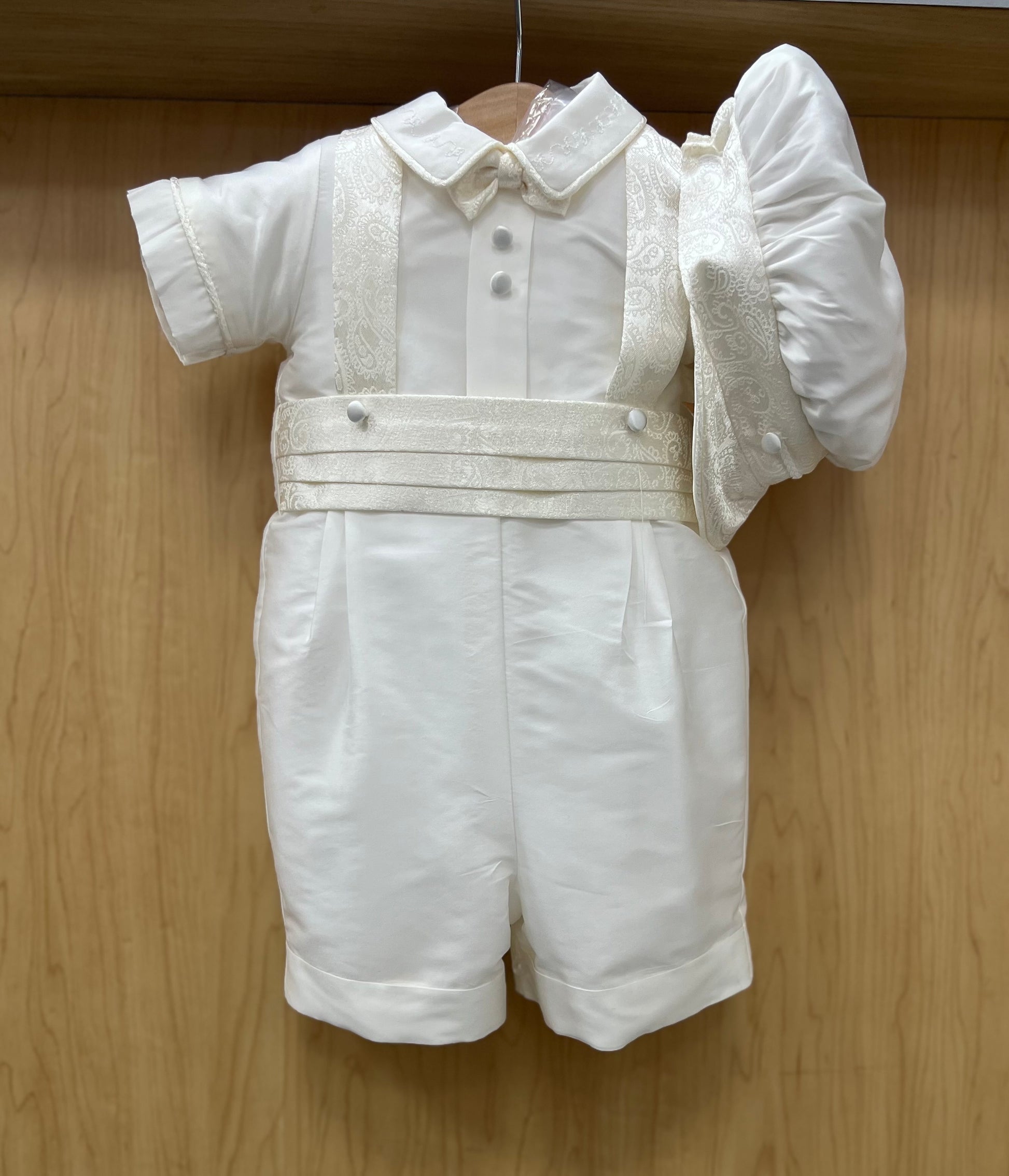 White silk baby shorts christening outfit with jacquard suspenders and a flat cap, displayed on a wooden hanger.