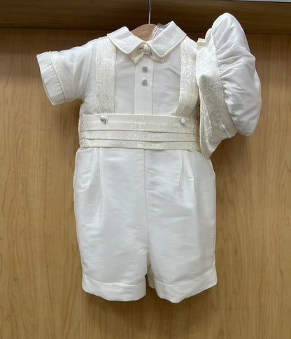 White silk baby shorts christening outfit with jacquard suspenders and a flat cap, displayed on a wooden hanger.