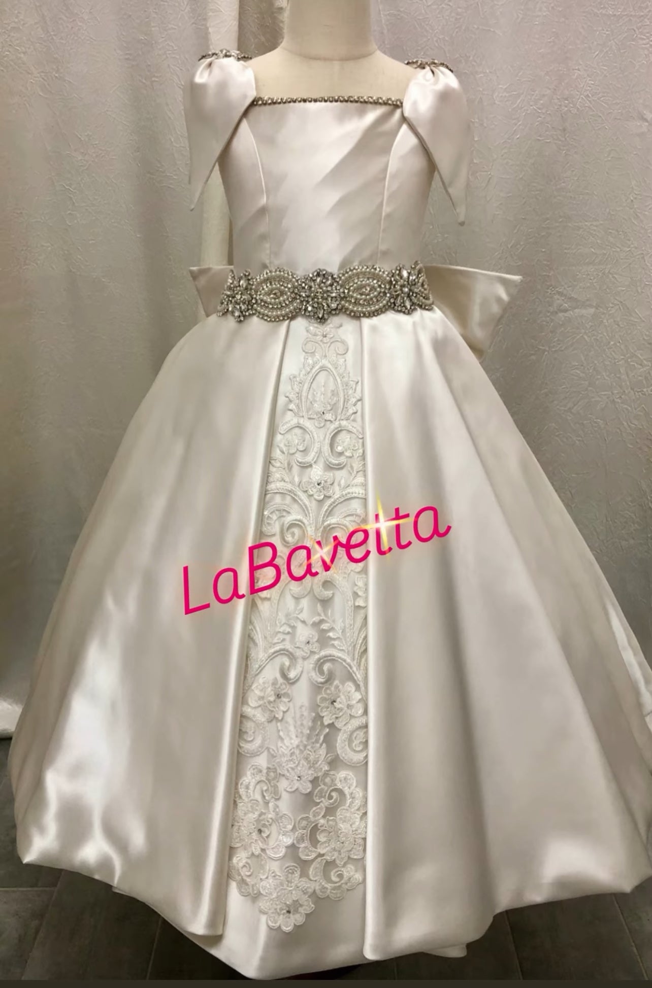 White satin communion dress with lace details and 'LaBavetta' branding on a neutral background